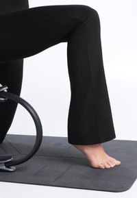 Person kneeling on a black exercise mat, wearing black pants, holding a black Pilates ring beside their bent knee.