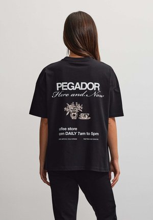 Persona con cabello largo de espaldas, llevando una camiseta negra con el texto "PEGADOR Here and Now,", taza de café y horarios de la tienda impresos en la parte trasera.