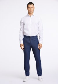 Weißes Hemd mit langen Ärmeln, offener Kragen, blaue Anzughose und weiße Sneaker. Schmale Passform mit glattem Stoff und dezenten Mustern auf dem Hosenstoff.