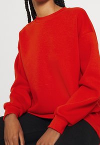 Röd sweatshirt med långa ärmar och ribbad rund halsringning. Materialet är mjukt med en slät yta. Har något sänkta axlar.