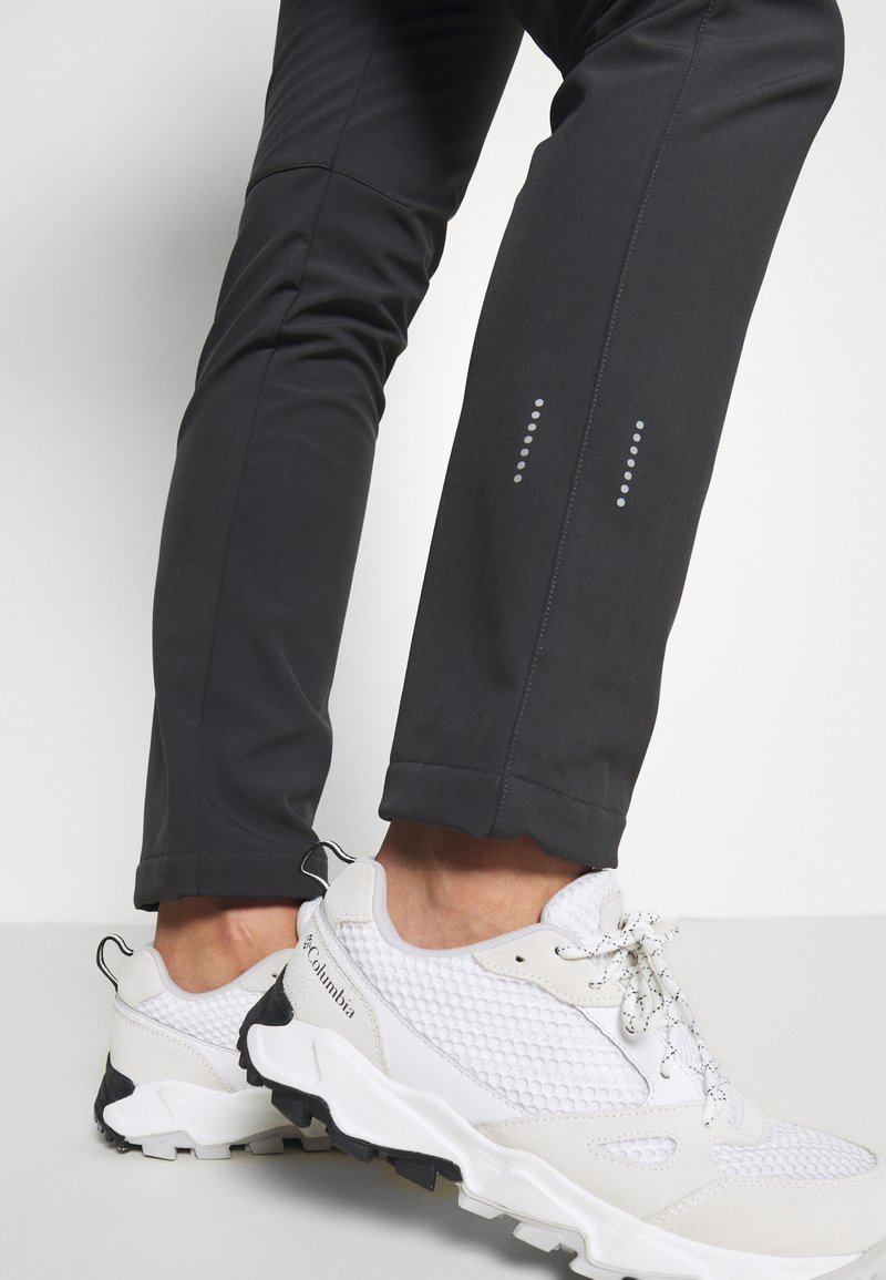 Zwarte, vochtafvoerende pantalons met reflecterende accenten en een taps toelopend ontwerp. Het model combineert ze met witte mesh sneakers met rubberen zolen.
