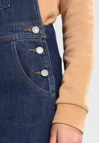 Boutons fermant une salopette en denim bleu foncé sur une personne portant une chemise beige à manches longues avec des poignets côtelés, la main reposant le long du corps.