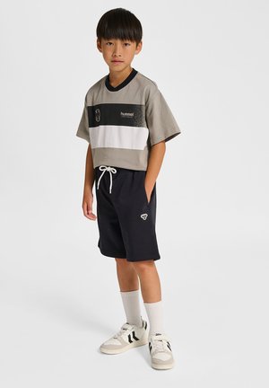 Junge trägt graues und schwarzes Colorblock-T-Shirt, schwarze Shorts mit Kordelzug, weiße Socken und graue Turnschuhe, steht mit einer Hand in der Tasche.