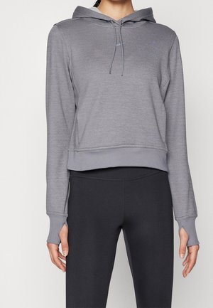 Femme portant un sweat à capuche gris avec des passages pour les pouces et un legging noir, debout devant un fond blanc uni.
