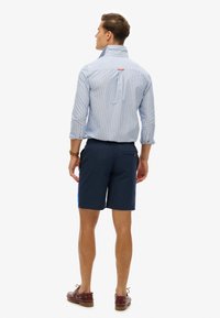 Chemise rayée bleu clair et blanche avec un col droit, portée avec un short marine. La vue de dos montre un détail de bouton caché au niveau du col.