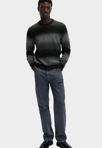 Gestrickter Pullover mit horizontalen Streifen in Dunkelgrün, Grau und Schwarz; Rundhalsausschnitt, gerippte Bündchen und Saum. Kombiniert mit grauen Jeans aus Denim.