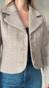 Žena v béžovém saku se vzorem rybí kost, s velkými klopami a třemi knoflíky, v kombinaci s modrými džíny s vysokým pasem.