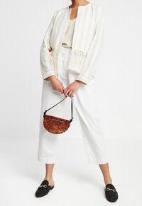 Ljusbeige och vitrandig jacka med fransar vid midjan, kombinerad med vita vida byxor och svarta texturerade loafers. Håller en halvcirkelformad vävd väska.