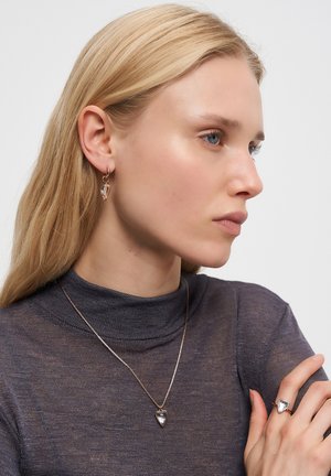 Blonde Frau in einem dunklen Rollkragenpullover, mit herzförmigen Kristallohrringen, passender Anhängerhalskette und Ring, die nach rechts schaut.