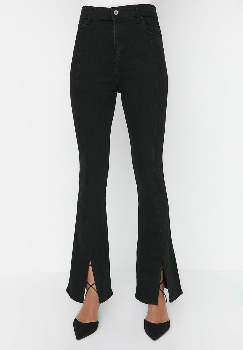 Pantalon noir taille haute slim avec fentes devant sur les jambes, porté avec des talons noirs pointus.