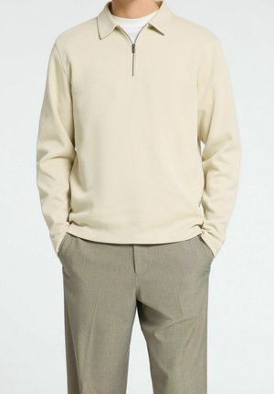 Homme portant une chemise beige clair à manches longues avec col zippé et un pantalon gris, les mains dans les poches, sur un fond blanc uni.