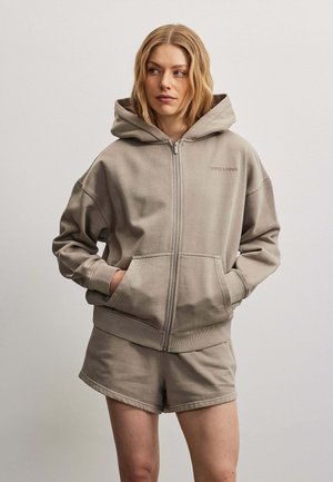 Donna che indossa una felpa con cappuccio beige con cerniera, con le mani nelle tasche e pantaloncini abbinati, in piedi davanti a uno sfondo chiaro e uniforme.