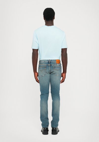 Light blue cotton t-shirt paired with fitted, mid-blue denim jeans. Back view shows a tan leather patch at the waistband and regular stitching.