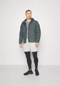 Grüne Daunenjacke mit Reißverschluss vorne, Kapuze und kontrastierenden schwarzen Akzenten. Darunter ein graues Oberteil, cremefarbene Shorts und schwarze Leggings, kombiniert mit schwarzen Schuhen.
