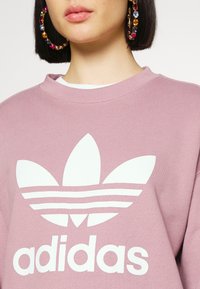 Persona con sudadera Adidas color malva con un gran logo blanco y pendientes de aro con piedras preciosas de colores, recortada a la altura del mentón y los hombros.