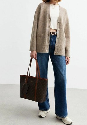 Personne portant un cardigan beige boutonné, un top court blanc, un jean bleu et des baskets blanches tenant un sac cabas marron foncé avec de longues anses.