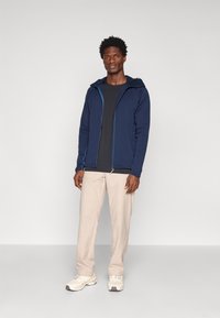 Tumesinine fliisjakki kapuutsiga, millel on kontrastsed sinised detailid. Kannan musta särgi peal, koos heledate beežide pükste ja heledate tossudega.