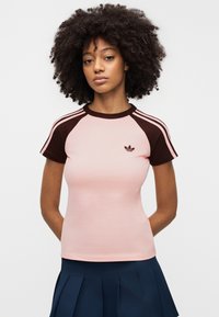 Różowy dopasowany t-shirt z brązowymi rękawami raglanowymi i trzema białymi paskami na każdym barku. Na piersi znajduje się małe czarne logo.