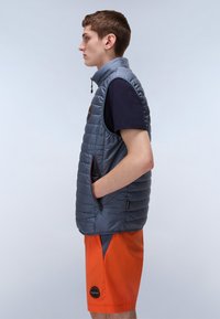 Padded gray vest with high collar and zip pockets. Worn over a navy shirt and paired with orange shorts, featuring a logo on the left leg.