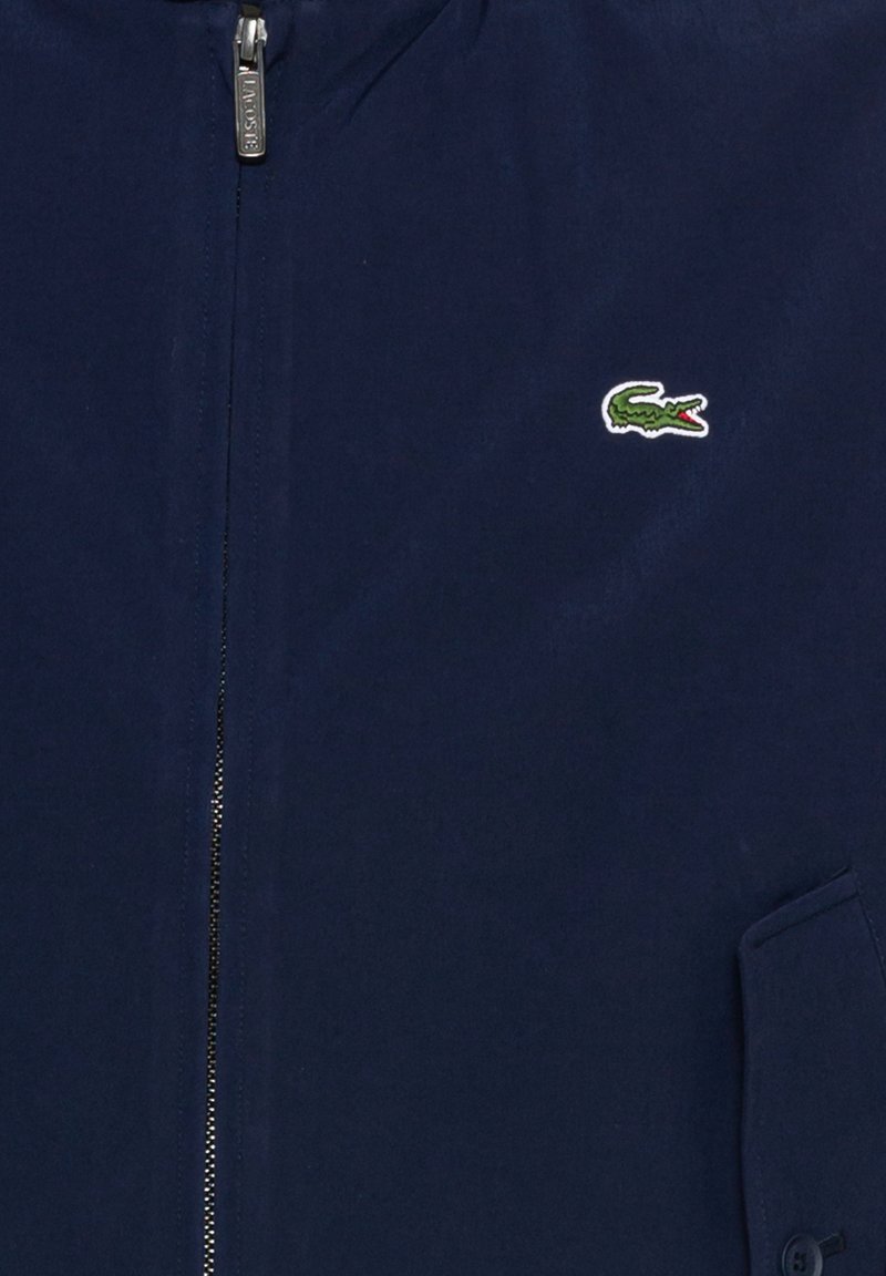 Veste zippée bleu marine en texture lisse, arborant un petit logo crocodile vert et blanc sur la poitrine gauche et une poche latérale.