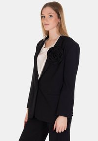 Blazer nero con grande accentuazione in tessuto a forma di rosa sulla spalla sinistra, caratterizzato da un design con revers classico e due tasche frontali. Texture morbida.