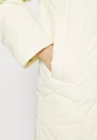 Mano dentro del bolsillo derecho de una chaqueta acolchada color crema con mangas largas y un sutil diseño geométrico en la costura.