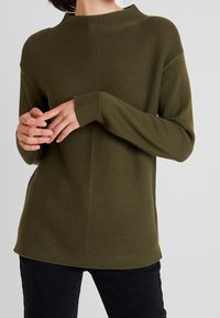 Olivgrüner gestrickter Pullover mit hohem Kragen, langen Ärmeln, strukturiertem Muster und gerippten Bündchen. Dezentes Markenlogo an der Seite.