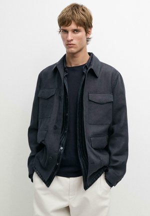 Junger Mann mit kurzem hellbraunem Haar, der eine dunkle, zugeknöpfte Jacke über einem schwarzen Hemd und cremefarbenen Hosen trägt, vor einem schlichten Hintergrund stehend.