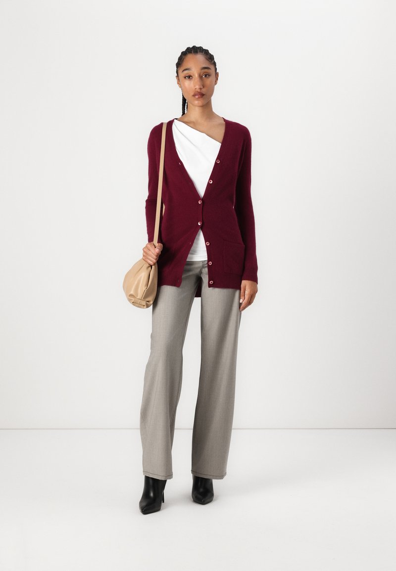 Burgunderfarbener Cardigan mit Knöpfen und Taschen, kombiniert mit einem weißen Oberteil, hellgrauen Hosen und schwarzen Absatzstiefeletten. Tanfarbene Schultertasche.