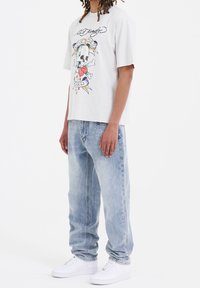 Camiseta gráfica de algodón gris claro, con un diseño colorido de calavera y tibias cruzadas. Combinada con unos pantalones vaqueros de mezclilla ligera y holgados en azul claro.