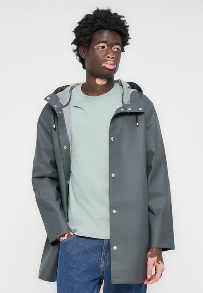 Vannavvisende grå jakke med snøring i hetta, sølvknapper og lysegrå fòr, båret over en lys grønn skjorte og jeans.