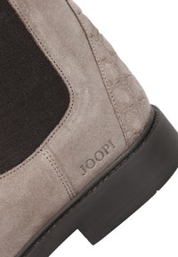 Botín de ante en gris claro con paneles laterales elásticos negros. Presenta un patrón texturizado y una suela de goma negra. Logotipo "JOOP!" grabado.
