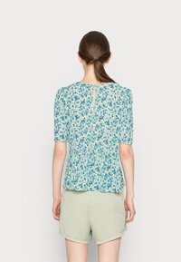 Blusa floral com padrões azuis e verdes, mangas curtas bufantes, decote redondo e detalhe de fenda nas costas. Combinada com calções verdes claros.