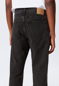 Jeans in denim nero con taglio a gamba dritta, caratterizzati da due tasche sul retro e un'etichetta in pelle marrone con la scritta "WEEKDAY" sulla cintura.