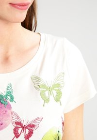 Off-white t-shirt med broderi av färgglada fjärilar i grön, röd och blå, med rund halsringning och korta ärmar.