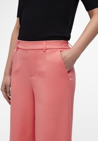 Pantalones rosas ajustados con una textura suave, bolsillos laterales y presillas para cinturón, combinados con una blusa negra de canalé ajustada.