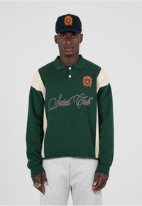 Polo a maniche lunghe verde e beige con colletto, logo ricamato e scritta "Social Club". Materiale in cotone, vestibilità rilassata, polsini a costine.