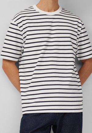 Mand iført en hvid kortærmet T-shirt med sorte vandrette striber og mørkeblå jeans, stående med hænderne bag ryggen.