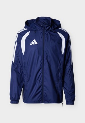 Tumesinise kapuutsiga tuulekindel jope valge kaarjate triipudega õlgadel ja ülemistel varrukatel, ees lukk ja elastsed mansetid.