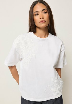 Junge Frau mit langen dunklen Haaren trägt ein weißes Kurzarmoberteil mit erhabener Blumenstickerei an der Schulter und an der unteren Seite.