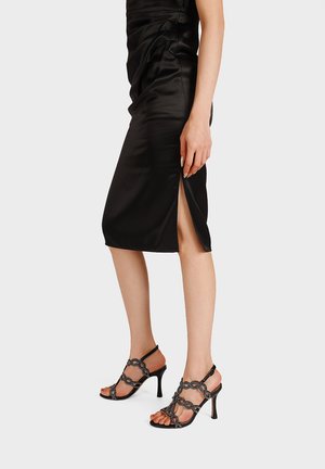 Donna indossa un abito midi in satin nero con spacco laterale e sandali neri con tacco alto e cinturini su sfondo semplice.