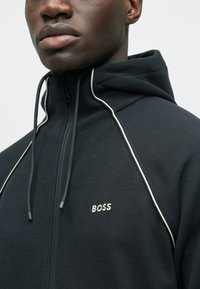 Μαύρο φούτερ με φερμουάρ, υφασμάτινη υφή, λευκές λεπτομέρειες κατά μήκος των ραφών και κεντημένο λογότυπο "BOSS" στο αριστερό στήθος.