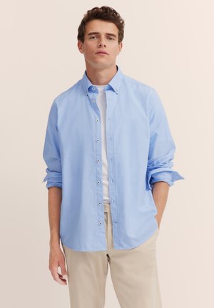 Junger Mann mit lockigem Haar, der ein hellblaues Button-Down-Hemd über einem weißen T-Shirt und beigen Hosen trägt, steht vor einem schlichten beigen Hintergrund.