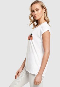 Hvid T-shirt med korte ærmer og rund hals, prydet med et grafik af et palmetræ over et solnedgangsmotiv i koral og sort. Glat tekstur.