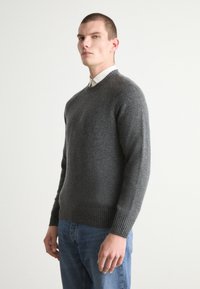 Maglione grigio lavorato a maglia con scollo a girocollo, polsini e orlo a costine. Indossato sopra una camicia con colletto e jeans blu. Texture morbida.