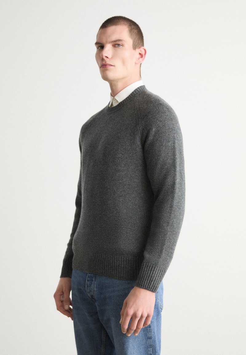 Maglione grigio lavorato a maglia con scollo a girocollo, polsini e orlo a costine. Indossato sopra una camicia con colletto e jeans blu. Texture morbida.