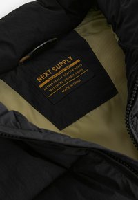Interno di giacca nera con foderatura khaki. Presenta un'etichetta rettangolare con la scritta "NEXT SUPPLY" e istruzioni per la cura ricamate in giallo.
