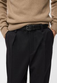 Pantalones negros de corte ajustado, combinados con un cinturón negro texturizado que presenta una hebilla redonda de plata, usados sobre un suéter de punto beige.