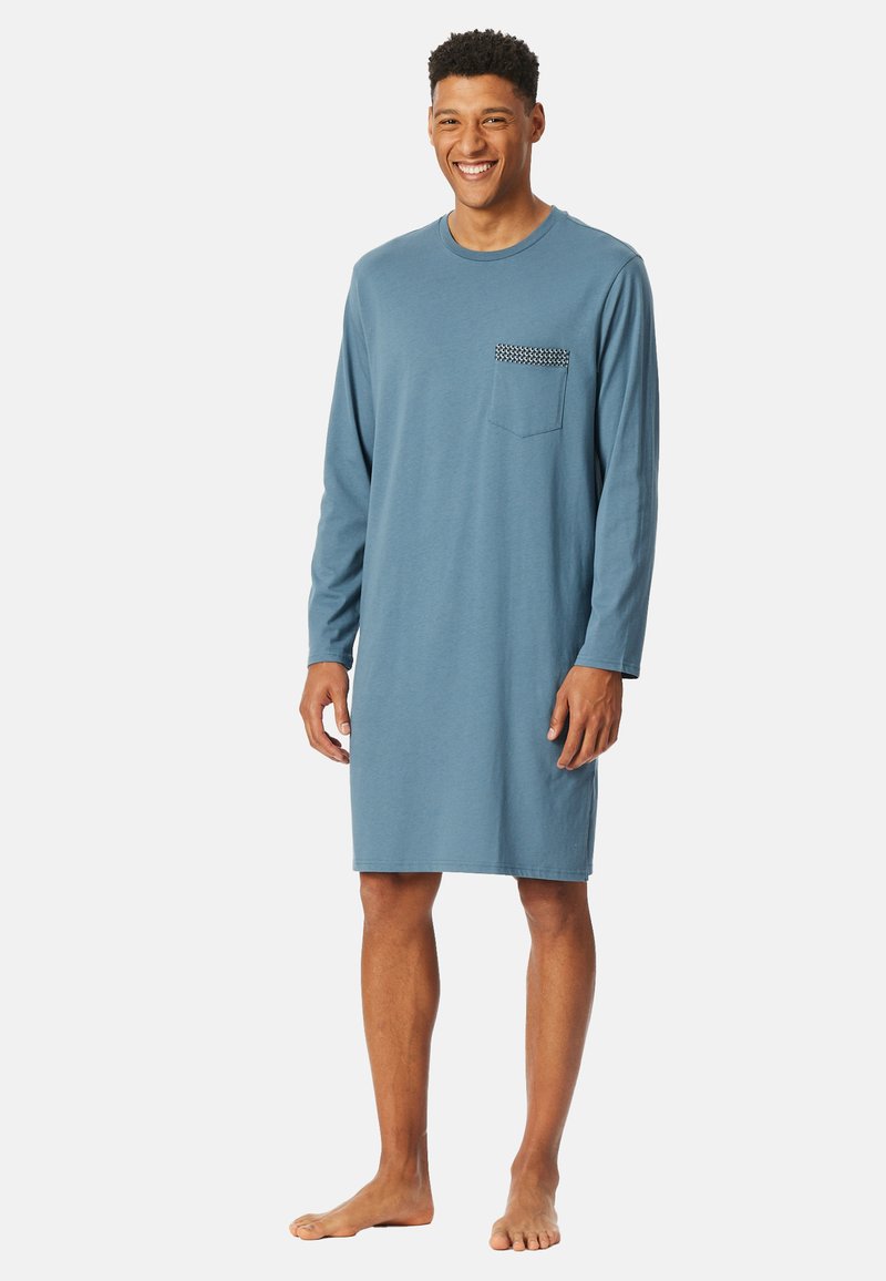 Homme souriant debout pieds nus, portant une chemise de nuit bleu à manches longues et arrivant jusqu'aux genoux avec une poche poitrine à motifs, sur fond blanc.