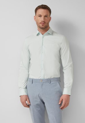 Mann trägt ein hellblaues, geknöpftes Hemd, das in hellgraue Anzughosen gesteckt ist, und steht vor einem schlichten, hellen Hintergrund.